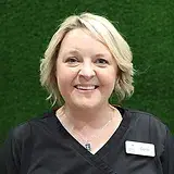 Professional headshot of a smiling woman with blonde hair wearing a black top against a green background.