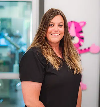 Woman in black shirt smiling at camera in dental office with pink signage visible.
