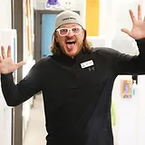 Enthusiastic dental professional wearing headwear and name badge, waving in welcoming gesture inside clinic.