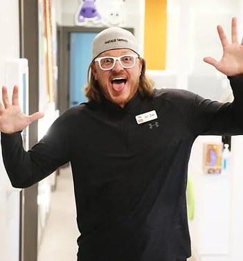 Enthusiastic dental staff member wearing beanie and glasses with arms raised in welcoming pose inside dental office.
