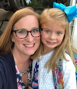 Two smiling females posing together in a car, one wearing glasses and a blue bow.