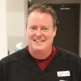Man in dark shirt with name badge smiling at camera in dental office setting.