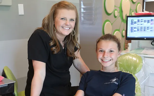 Two people smile in a colorful pediatric dental office with computer monitors visible in background.