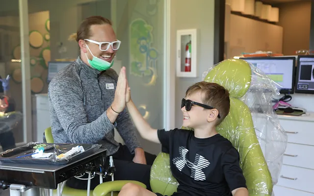 Pediatric dentist in glasses discusses treatment with young patient in dental office chair.