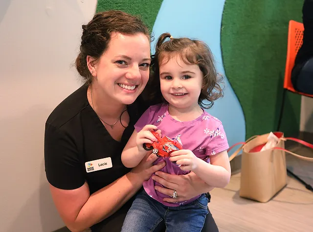 Pediatric dentist in black uniform smiles with young child holding toothbrush in colorful dental office.