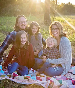 Family of five smiling together outdoors on blanket during golden hour sunset.